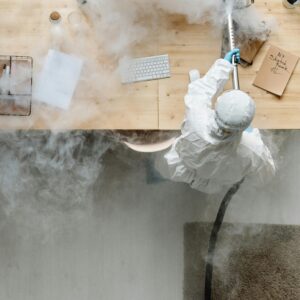 A person in protective gear sanitizing an office desk using fumigation equipment during daytime.
